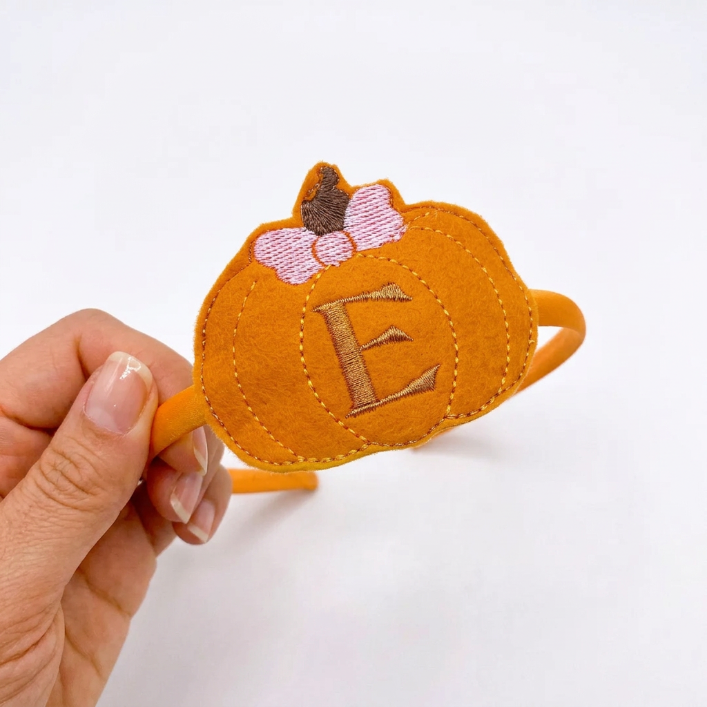 A hand holding a orange pumpkin-shaped feltie with the letter 'E' on it, intended as a hairband slider.